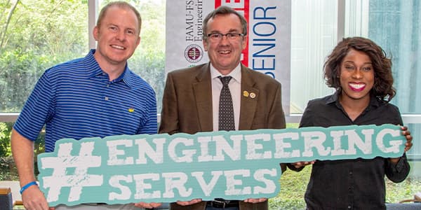 Tom Derzypolski, Dean Murray Gibson and Amber Smith stand together at the FAMU-FSU College of Engineering. The three of them hold up a sign that reads hashtag Engineering Serves, in blue text.